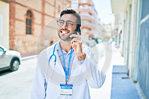 Young handsome hispanic doctor wearing stethoscope smiling happy Standing with smile on face talking on the smartphone at town