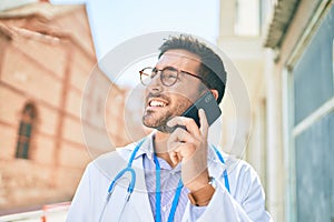 Young handsome hispanic doctor wearing stethoscope smiling happy Standing with smile on face talking on the smartphone at town