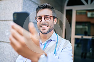 Young handsome hispanic doctor wearing uniform and stethoscope smiling happy Standing with smile on face using smartphone at town