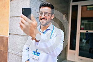 Young handsome hispanic doctor wearing uniform and stethoscope smiling happy Standing with smile on face using smartphone at town