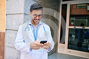 Young handsome hispanic doctor wearing uniform and stethoscope smiling happy Standing with smile on face using smartphone at town