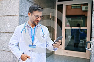 Young handsome hispanic doctor wearing uniform and stethoscope smiling happy Standing with smile on face using smartphone at town