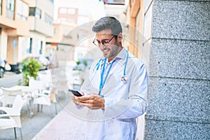 Young handsome hispanic doctor wearing uniform and stethoscope smiling happy Standing with smile on face using smartphone at town