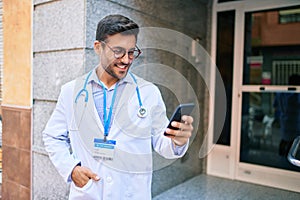 Young handsome hispanic doctor wearing uniform and stethoscope smiling happy Standing with smile on face using smartphone at town