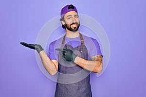 Young handsome hispanic man with bear wearing professional apron working as tattoo artist amazed and smiling to the camera while