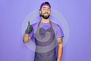 Young handsome hispanic man with bear wearing professional apron working as tattoo artist doing happy thumbs up gesture with hand