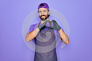 Young handsome hispanic man with bear wearing professional apron working as tattoo artist smiling in love showing heart symbol and