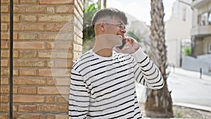 Young, handsome hispanic man confidently smiling, enjoying sunny day on city street, casually leaning against wall, talking on