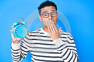 Young handsome hispanic man holding alarm clock covering mouth with hand, shocked and afraid for mistake