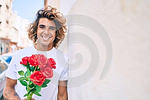 Young handsome hispanic man holding bouquet of roses leaning on the wall at street of city