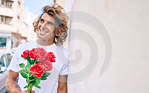 Young handsome hispanic man holding bouquet of roses leaning on the wall at street of city