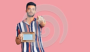 Young handsome hispanic man holding empty frame pointing with finger to the camera and to you, confident gesture looking serious