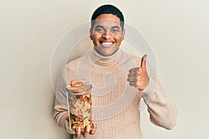 Young handsome hispanic man holding jar with farfalle pasta smiling happy and positive, thumb up doing excellent and approval sign