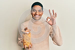 Young handsome hispanic man holding jar with macaroni pasta doing ok sign with fingers, smiling friendly gesturing excellent
