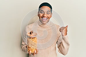 Young handsome hispanic man holding jar with macaroni pasta pointing thumb up to the side smiling happy with open mouth
