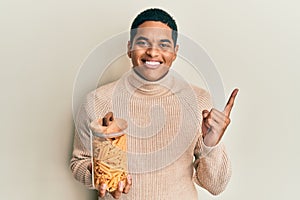 Young handsome hispanic man holding jar with macaroni pasta smiling happy pointing with hand and finger to the side