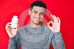 Young handsome hispanic man holding pills doing ok sign with fingers, smiling friendly gesturing excellent symbol