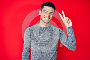 Young handsome hispanic man over red background smiling with happy face winking at the camera doing victory sign