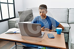 Young handsome hispanic man using laptop sitting on the floor with hand on stomach because indigestion, painful illness feeling