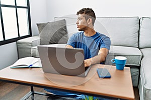 Young handsome hispanic man using laptop sitting on the floor looking to side, relax profile pose with natural face with confident