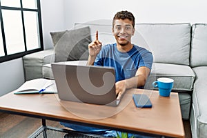 Young handsome hispanic man using laptop sitting on the floor showing and pointing up with finger number one while smiling