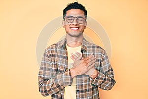 Young handsome hispanic man wearing casual clothes and glasses smiling with hands on chest with closed eyes and grateful gesture