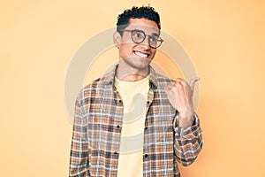 Young handsome hispanic man wearing casual clothes and glasses smiling with happy face looking and pointing to the side with thumb