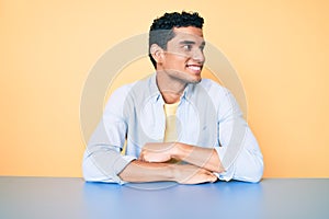 Young handsome hispanic man wearing casual clothes sitting on the table looking away to side with smile on face, natural