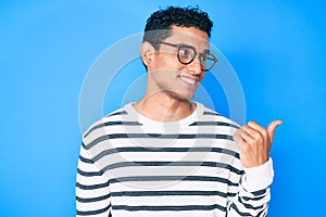 Young handsome hispanic man wearing casual sweater and glasses smiling with happy face looking and pointing to the side with thumb