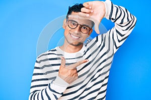 Young handsome hispanic man wearing casual sweater and glasses smiling making frame with hands and fingers with happy face