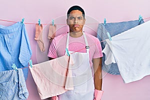 Young handsome hispanic man wearing cleaner apron holding clothes on clothesline depressed and worry for distress, crying angry