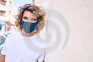 Young handsome hispanic man wearing coronavirus protection mask leaning on the wall at street of city