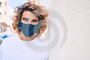 Young handsome hispanic man wearing coronavirus protection mask leaning on the wall at street of city