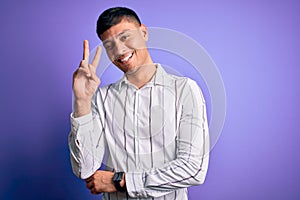 Young handsome hispanic man wearing elegant business shirt standing over purple background smiling with happy face winking at the