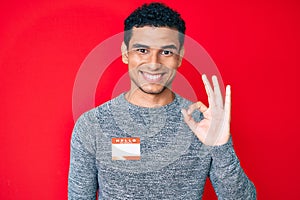 Young handsome hispanic man wearing hello my name is sticker identification doing ok sign with fingers, smiling friendly gesturing