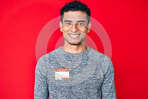 Young handsome hispanic man wearing hello my name is sticker identification looking positive and happy standing and smiling with a