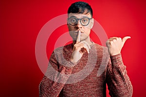 Young handsome hispanic man wearing nerd glasses over red background asking to be quiet with finger on lips pointing with hand to