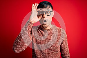 Young handsome hispanic man wearing nerd glasses over red background doing ok gesture shocked with surprised face, eye looking