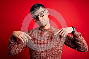 Young handsome hispanic man wearing nerd glasses over red background Pointing down looking sad and upset, indicating direction