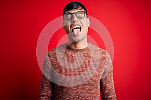 Young handsome hispanic man wearing nerd glasses over red background sticking tongue out happy with funny expression