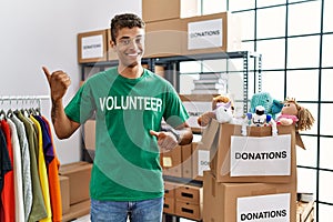 Young handsome hispanic man wearing volunteer t shirt at donations stand pointing to the back behind with hand and thumbs up,