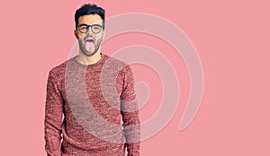 Young handsome hispanic man wearing winter sweater and glasses sticking tongue out happy with funny expression