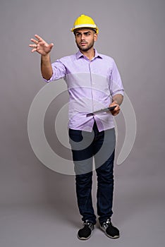 Young handsome Indian businessman wearing hardhat against gray b