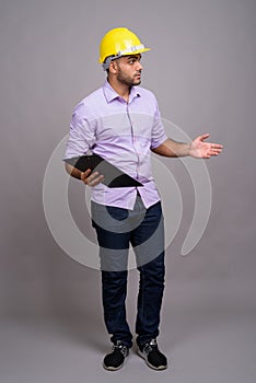 Young handsome Indian businessman wearing hardhat against gray b