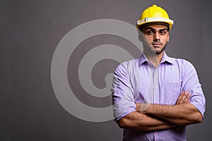 Young handsome Indian businessman wearing hardhat against gray b