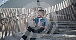 young handsome indian hindu man student freelancer sitting outdoors on steps using laptop computer