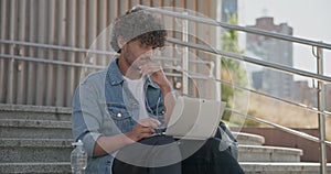 young handsome indian hindu man student freelancer sitting outdoors on steps using laptop computer