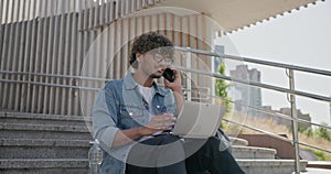 young handsome indian hindu man student freelancer sitting outdoors on steps using laptop computer and talking on the