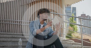 young handsome indian hindu man student freelancer sitting outdoors on steps using tablet pc computer pad