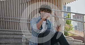 young handsome indian hindu man student freelancer sitting outdoors on steps using tablet pc computer pad